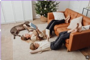Photo d'une mère et de sa fille en train de partager un moment de lecture. Elles sont allongées sur un tapis les jambes sur l'assise du canapé; un chien est allongé à côté d'elles. Ce moment de partage contribuer à donner envie de lire aux jeuens.